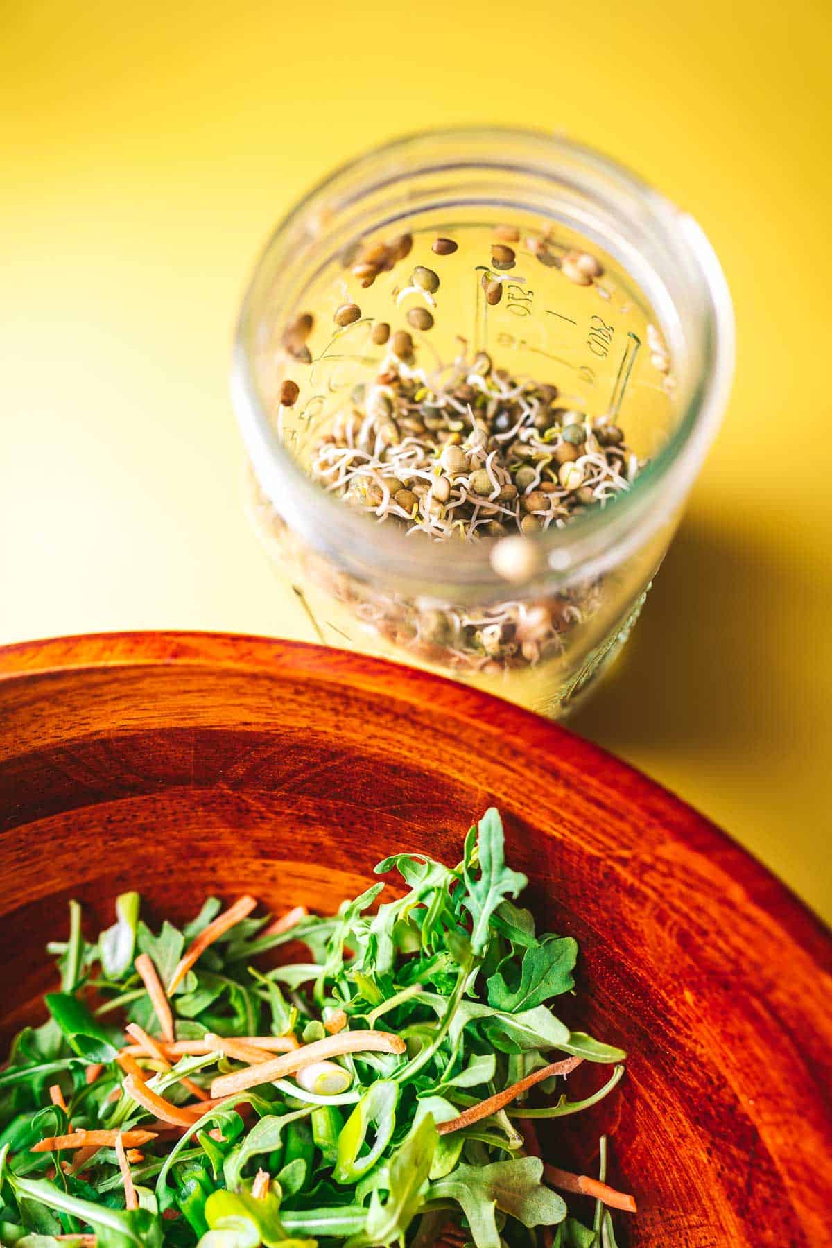 A jar of lentil sprouts resting on a yellow table next to a wooden salad bowl filled with green salad.