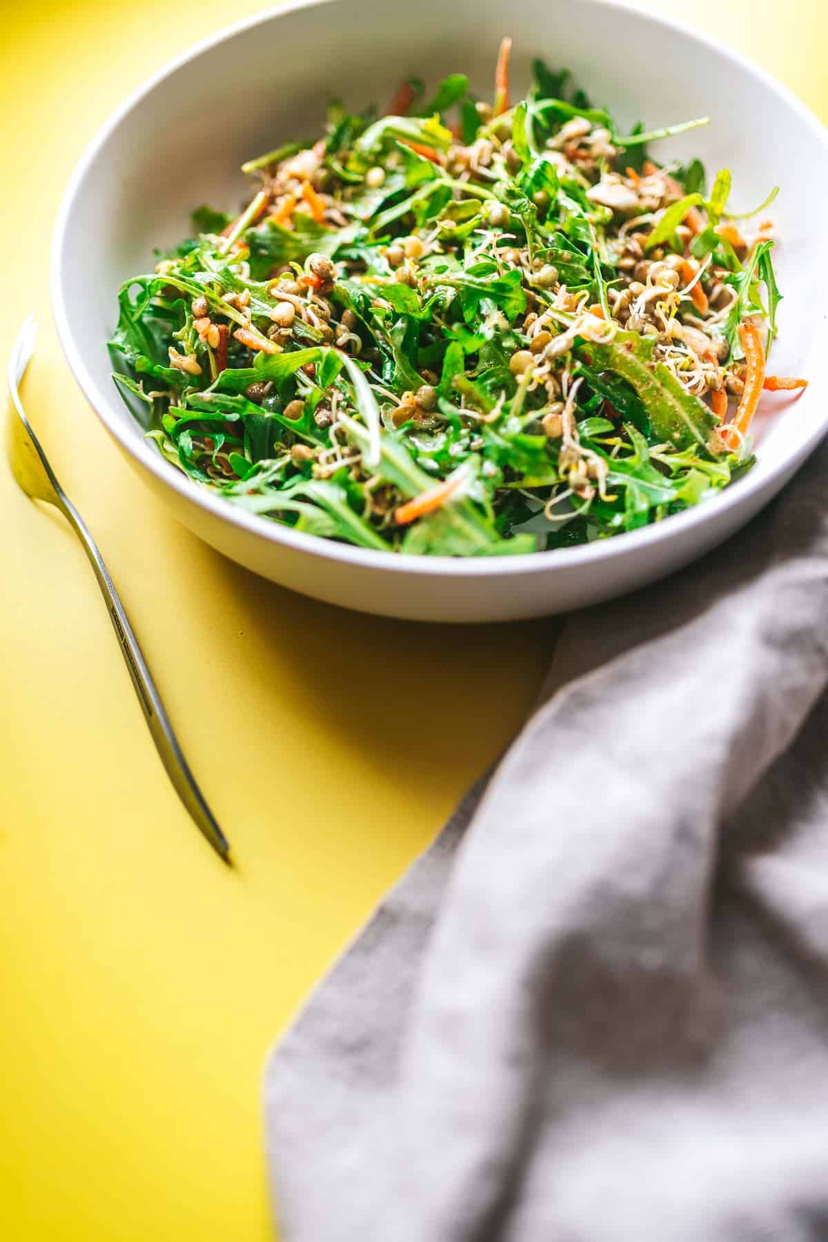 A white ceramic bowl filled with a sprouted lentil salad resting on a bright yellow tabletop.