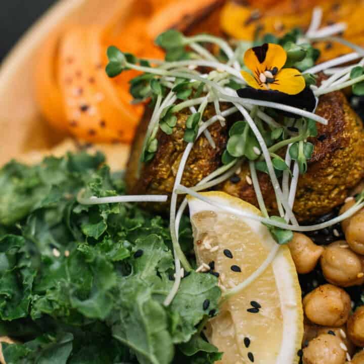 A close-up of a plant-based dish featuring chickpeas, leafy greens, sliced tomato, a lemon wedge, sprouts, and an edible flower.