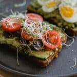 Two slices of toast topped with avocado, tomato, mushrooms, sprouts, and one with sliced boiled egg, served on a dark plate.