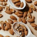 Whole almonds and walnut pieces, perfect for sprouting nuts and seeds, are scattered on a beige surface, with two wooden spoons holding some of the nuts.