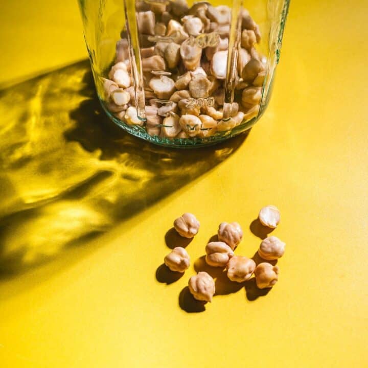 A glass jar filled with dried chickpeas sits on a yellow surface, with several chickpeas scattered outside the jar—perfect for demonstrating how to sprout chickpeas at home.