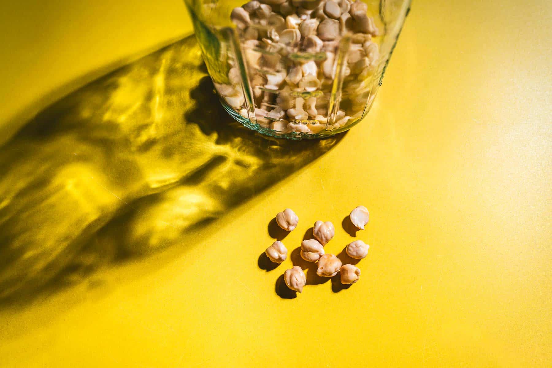 A glass jar filled with chickpeas sits on a yellow surface, with several chickpeas scattered outside the jar and a shadow cast to the left—perfect for learning how to sprout chickpeas at home.