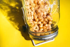 A glass jar filled with chickpeas, perfect for learning how to sprout chickpeas, lies on its side on a bright yellow surface, sunlight casting dramatic shadows and reflections.