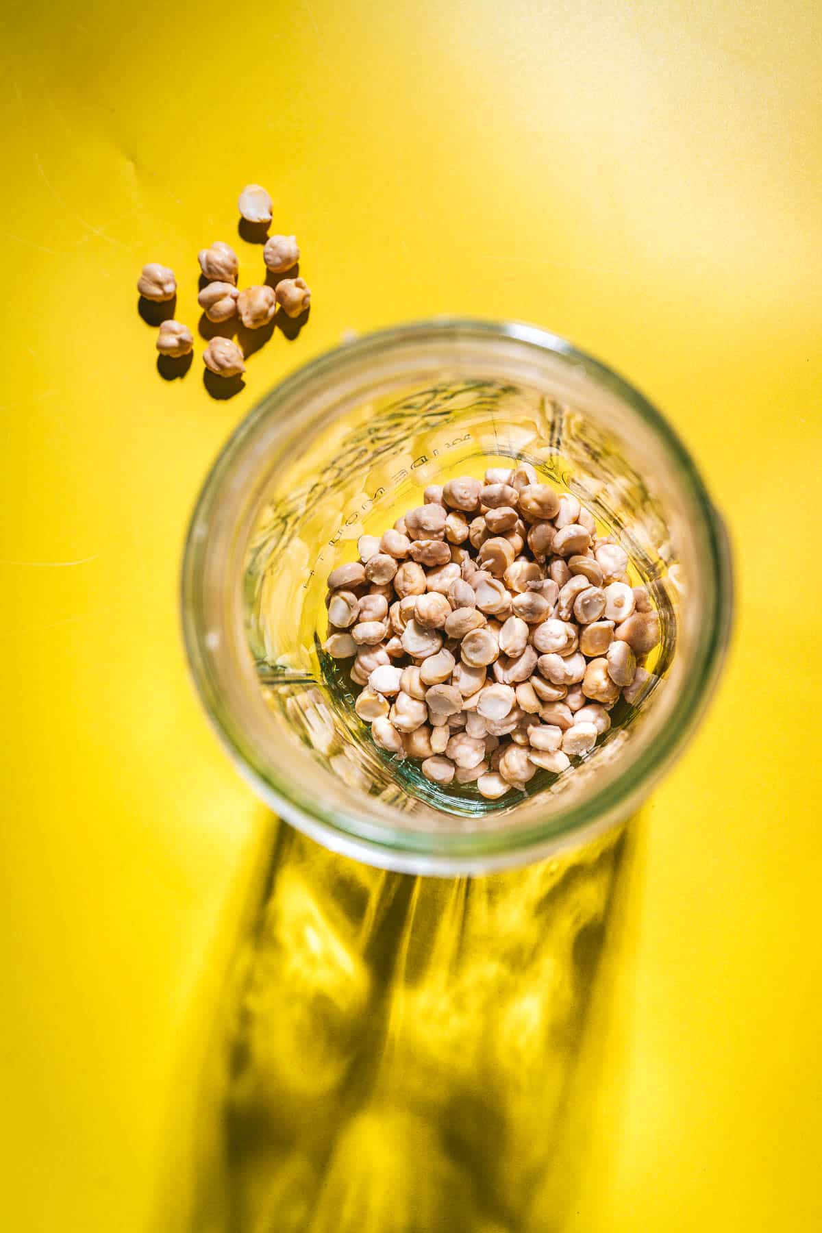 A glass jar with dried chickpeas inside sits on a yellow surface, several chickpeas scattered outside—perfect for learning how to sprout chickpeas at home.