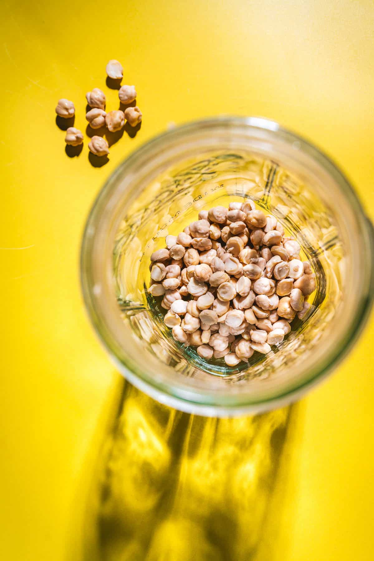A glass jar with chickpeas inside sits on a bright yellow surface, with a small pile of chickpeas next to it—perfect for showing how to sprout chickpeas at home.