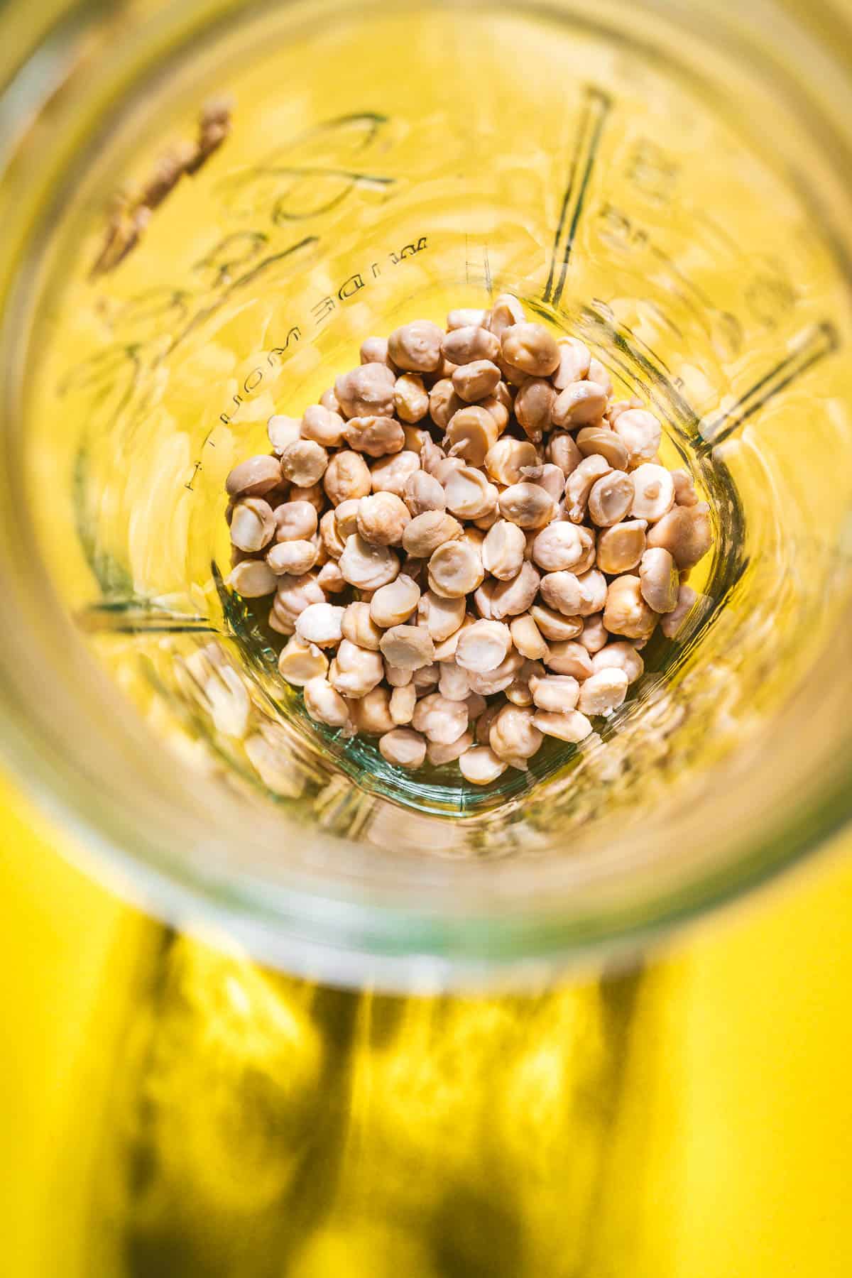 A glass jar containing a pile of chickpeas sits on a bright yellow surface, photographed from above—perfect for illustrating how to sprout chickpeas at home.