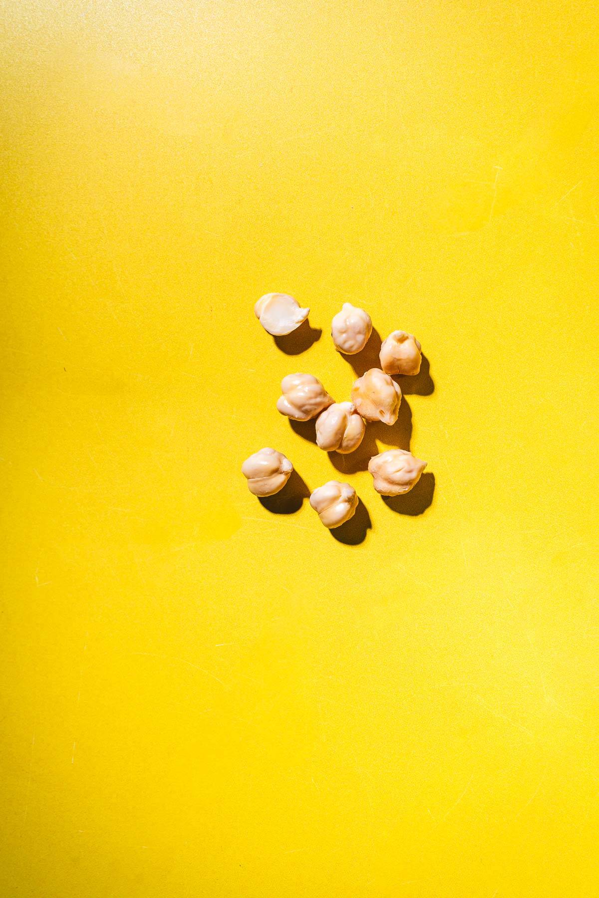 Nine chickpeas, perfect for learning how to sprout chickpeas, are scattered on a bright yellow surface, each casting a small shadow.
