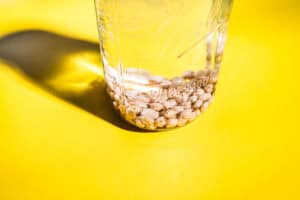 A glass jar filled with water and dried chickpeas, perfect for learning how to sprout chickpeas, sits on a bright yellow surface, casting a shadow.